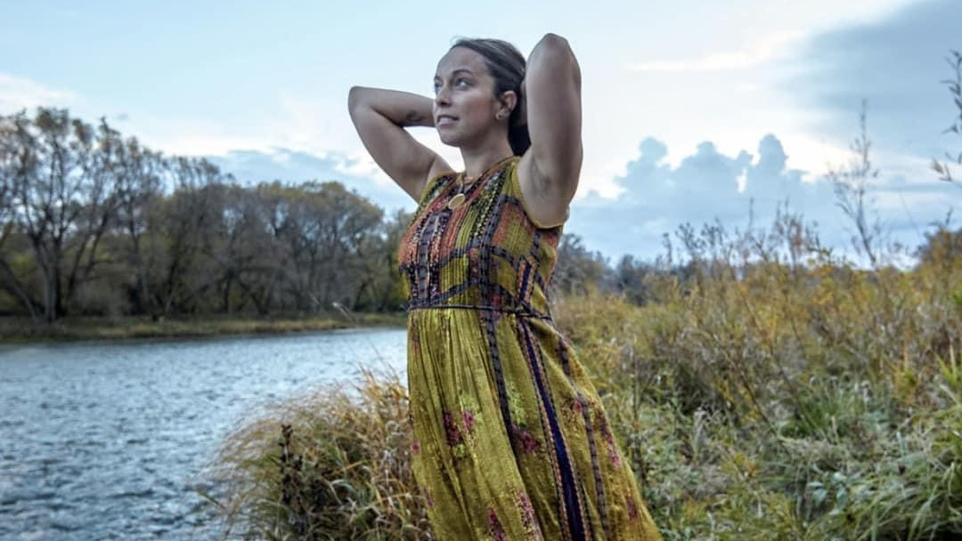 Evi Spiliotopoulos in an olive dress displaying wholehearted living while standing on the side of a river staring up at a cloud filled sky