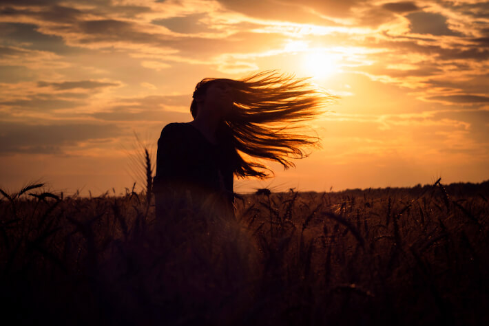 A woman stands in a field of wheat at sunset as the gold skyline stretches across the sky.