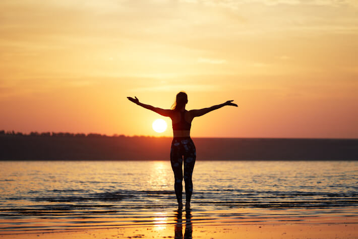 A woman dressed in activewear meant for Yoga, stands on the shore of a large body of water with her arms spread open wide.